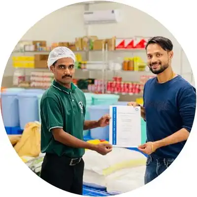 Two men are standing in a room filled with various items and boxes. The man on the left, wearing a green shirt and a hairnet, is holding a certificate. The man on the right, dressed in casual attire, is also holding the same certificate, smiling at the camera.