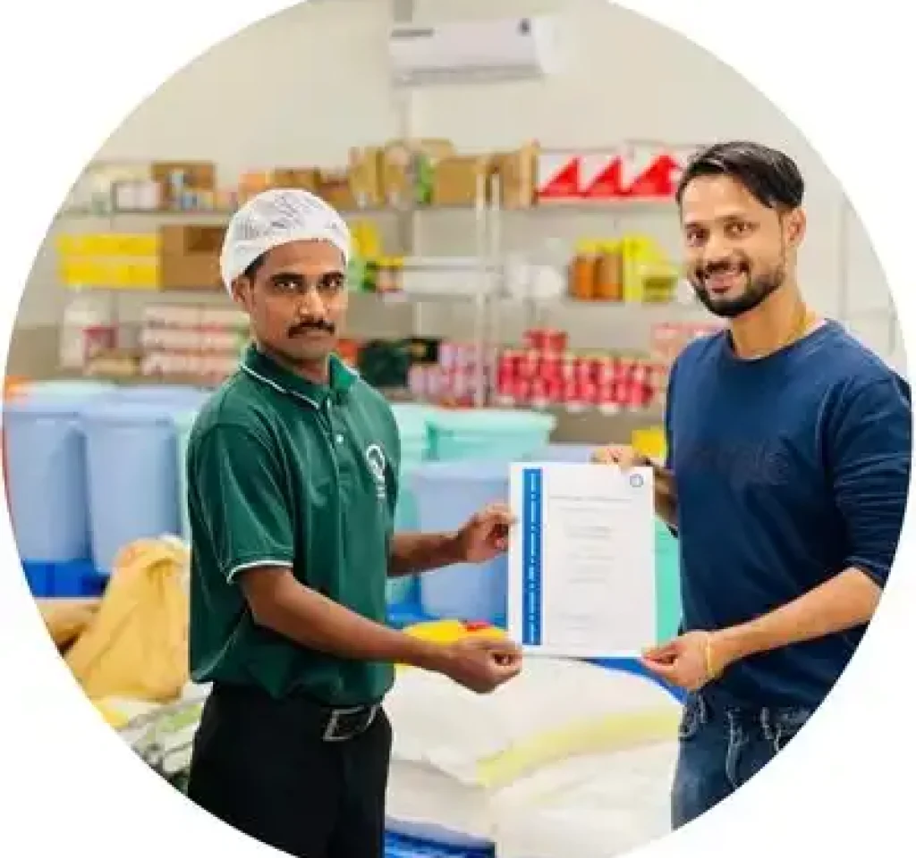 Two men are standing in a room filled with various items and boxes. The man on the left, wearing a green shirt and a hairnet, is holding a certificate. The man on the right, dressed in casual attire, is also holding the same certificate, smiling at the camera.